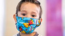 a Latino boy is wearing a teal colored facemask with cartoon characters in bright yellow and red